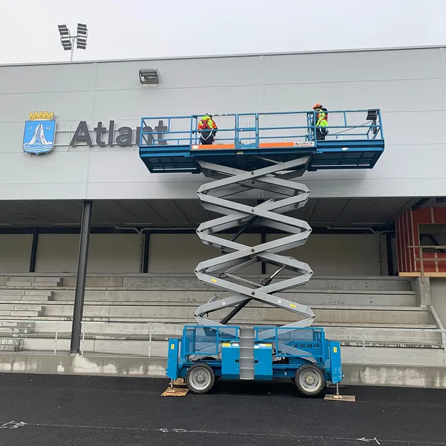 To personer på sakselift monterer skilt på grå tribunevegg med Atlant-logo.