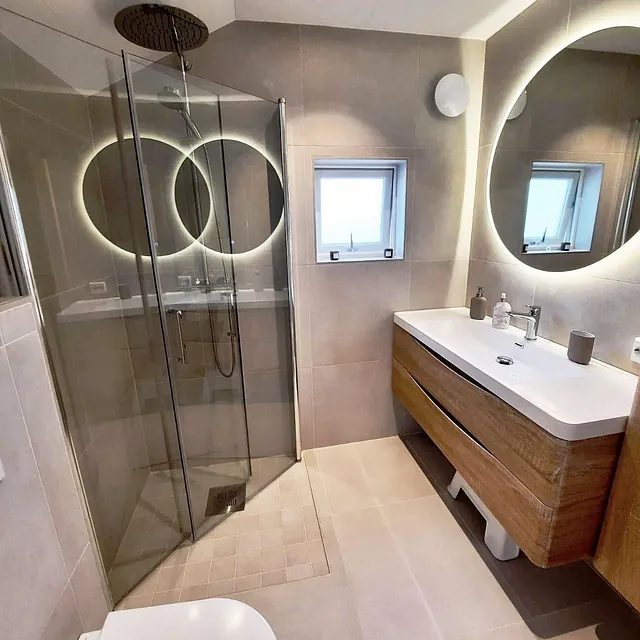 Lyst bad med dusjkabinett, treinnredning og rundt speil med lys.