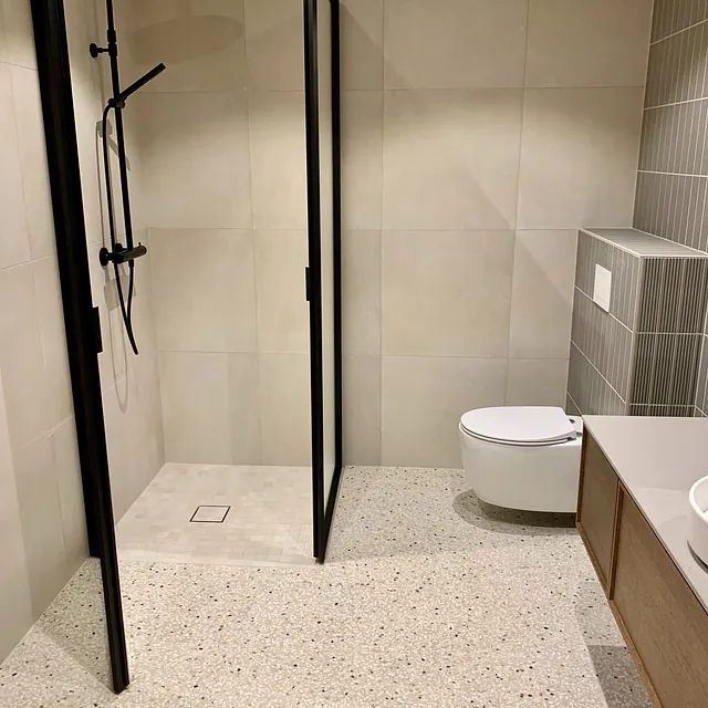 Bad med terrazzo-gulv, grå vegger, sort dusjarmatur og vegghengt toalett.