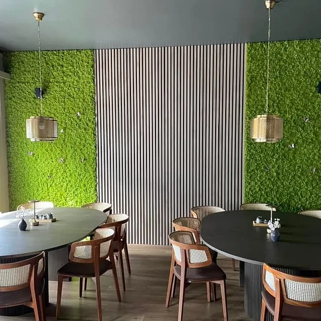 Rundt spisebord med tremøbler foran grønn mosevegg og hengende lamper.