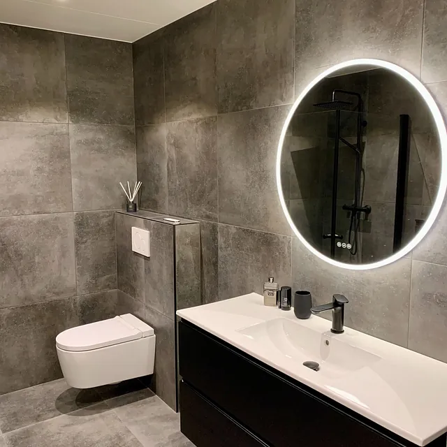 Bad med mørkegrå fliser, rundt lysende speil og svart servantinnredning.