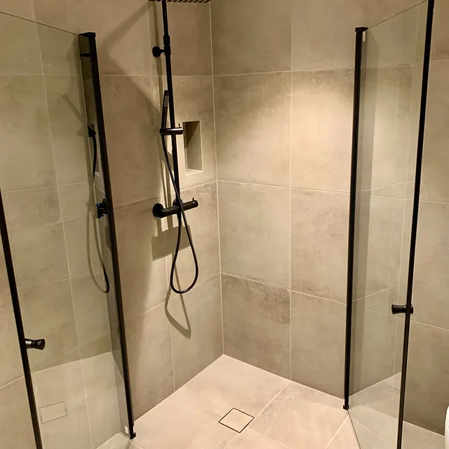 Dusjhjørne med store beige fliser, sorte armaturer og nisjehylle.