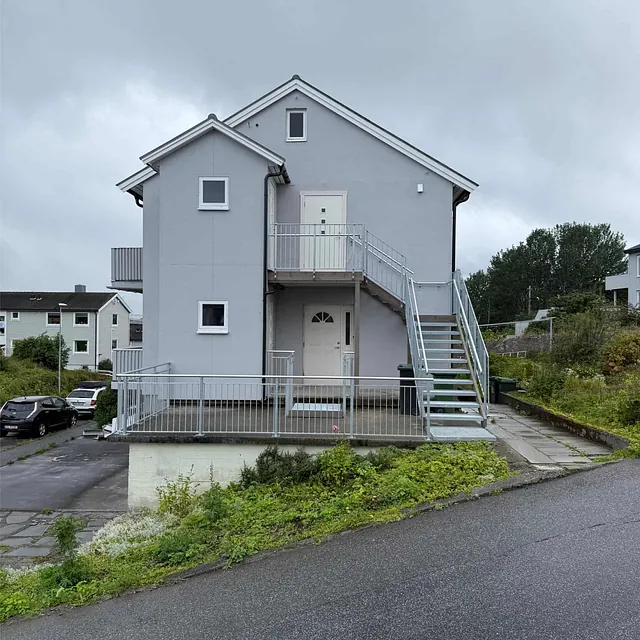 Hvit bolig med utvendig trapp og inngangsparti på hjørnetomt i stille nabolag.
