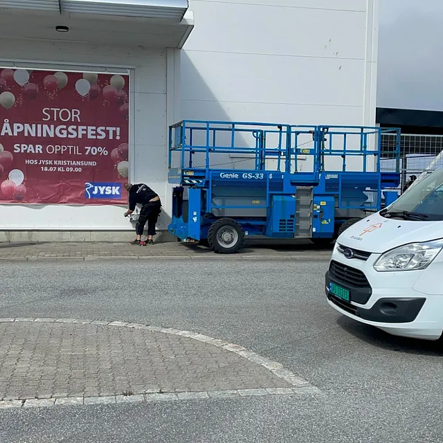 Arbeidere monterer plakat for JYSK åpningsfest, bil og lift foran.