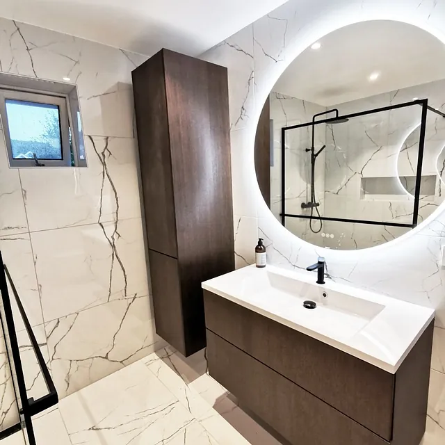 Moderne bad med marmorfliser, mørke skap og rundt speil med lys.