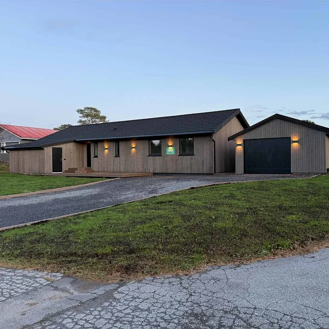 Moderne enebolig med trekledning og garasje plassert i skrånende terreng ved vei.