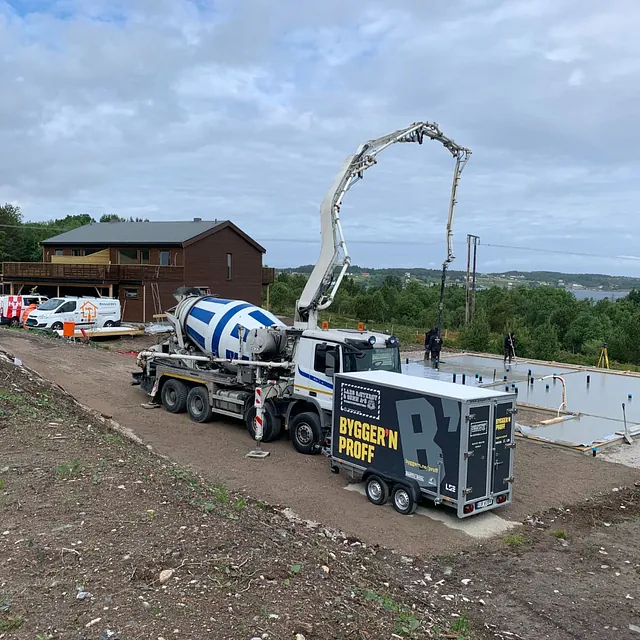 Betongbil støper grunnmur ved hus under bygging, flere firmabiler på stedet.