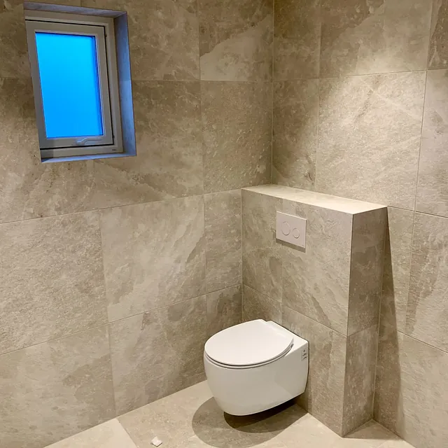 Bad med grå natursteinfliser, lite vindu og vegghengt toalett.