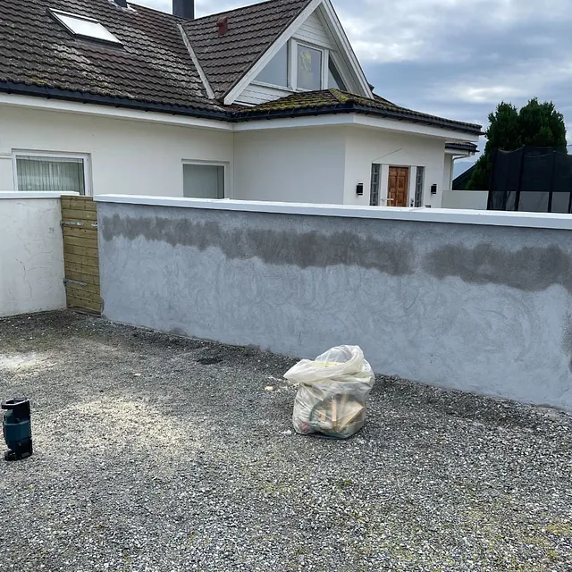 Delvis pusset mur foran hvitt hus med garasje og hage.