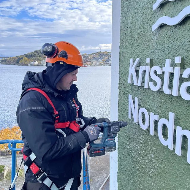 Arbeider med hjelm og drill monterer bokstaver på grønn husfasade ved sjøen.