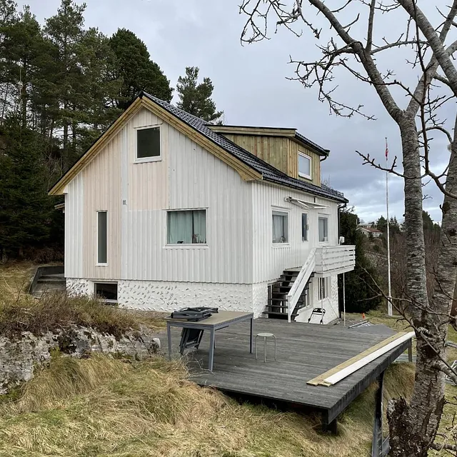 Hvit trehus med svart tak og tilbygg, sett fra hagen med stor terrasse.