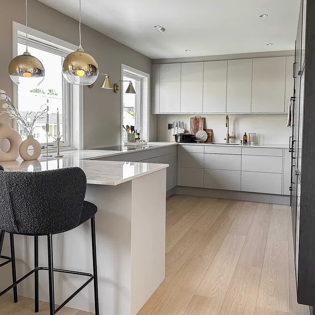 Moderne kjøkken med kjøkkenøy, lyse fronter, gullarmatur og store vinduer som slipper inn lys.