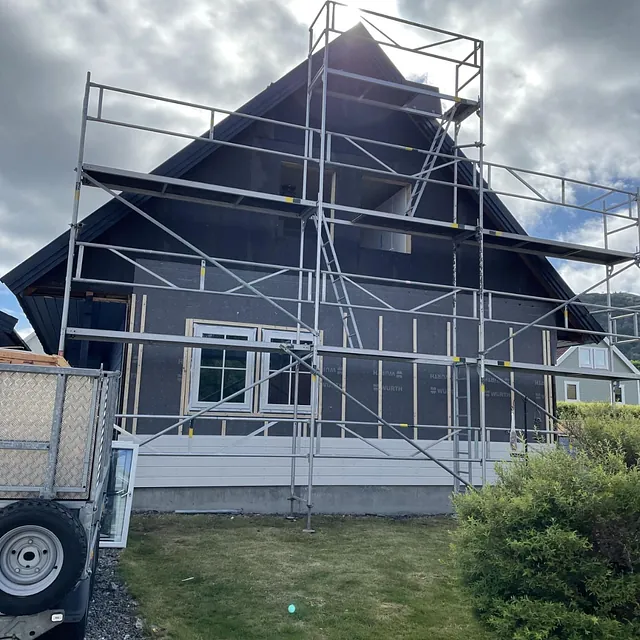 Hus med stillas under oppføring av ny kledning, grå plater synlige.