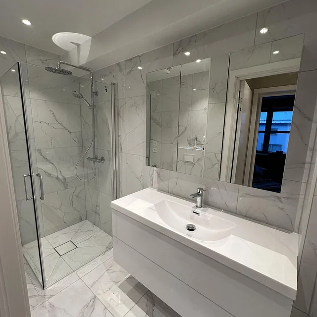 Lyst bad med hvite marmorfliser, dusjhjørne og hvit servant.