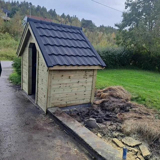 Liten trebod med saltak og svart takstein plassert ved en gressplen.