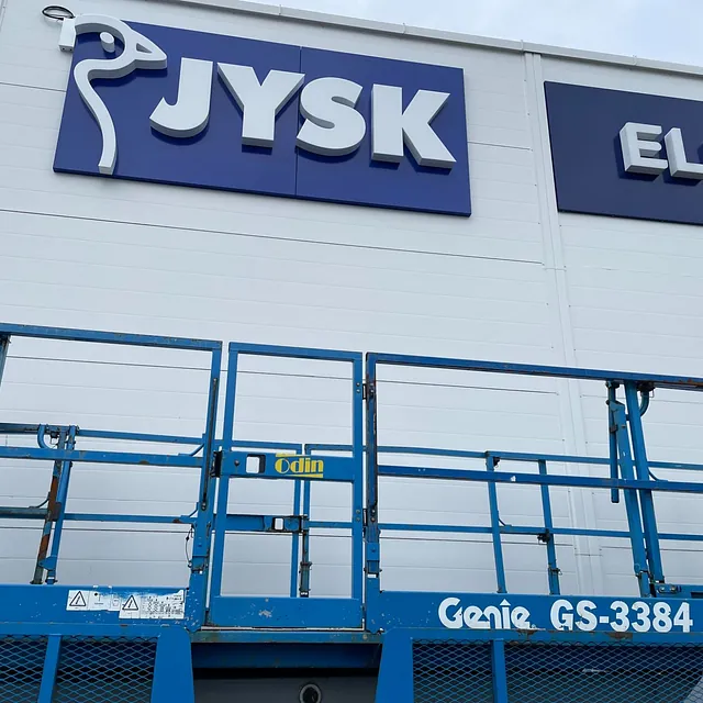 Arbeidere på lift foran bygg med JYSK- og Elkjøp-skilt på veggen.
