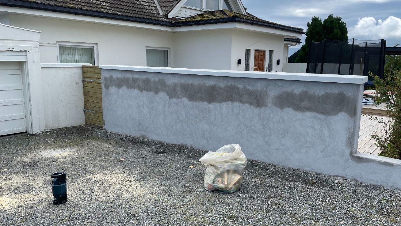 Hvit bolig med ny murvegg i grå puss foran gårdsplass.