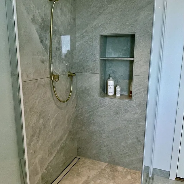 Dusj med grå steinfliser, gullfarget dusjarmatur og nisjehyller.