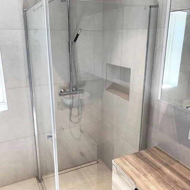 Lys dusj med store fliser, glassdører og trebenk ved siden.