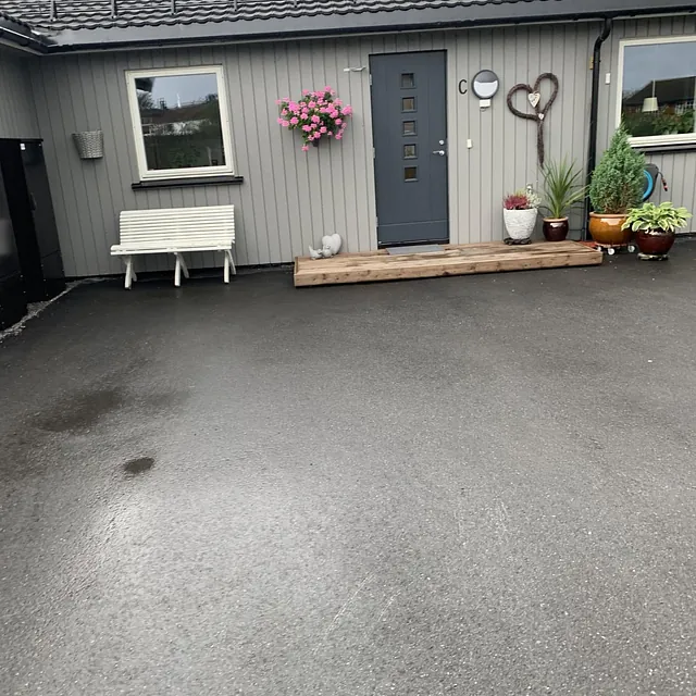 Grå enebolig med mørk dør, blomsterkrukker og benk ved inngangspartiet.