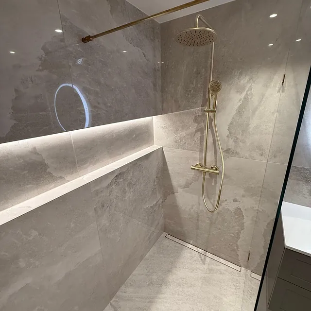 Romslig dusjsone med store fliser, innebygd hylle og moderne armatur i gull.