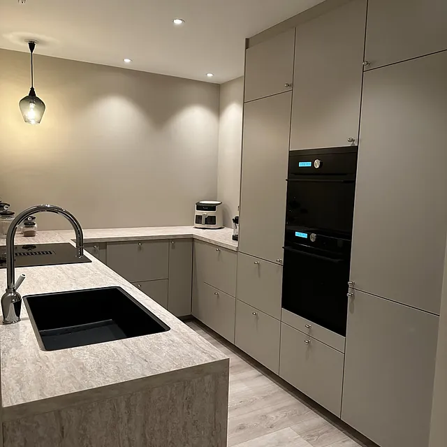 Moderne kjøkken med beige fronter, sorte hvitevarer og lys steinbenk.