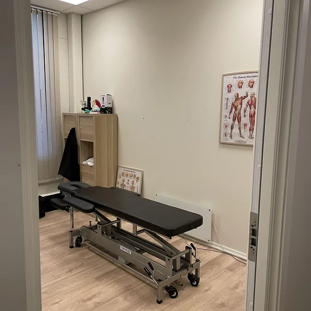 Behandlingsrom med svart benk, anatomiplakat og skap mot veggen.