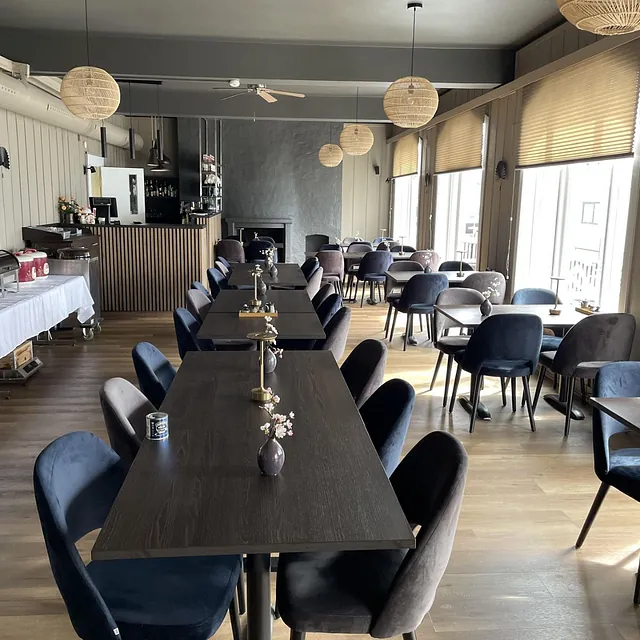 Restaurant med mørke bord og stoler, peis og bar i enden av rommet.