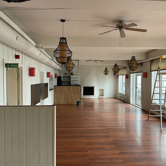 Stort tomt rom med trelaminatgulv, peis og vinduer på langsiden.