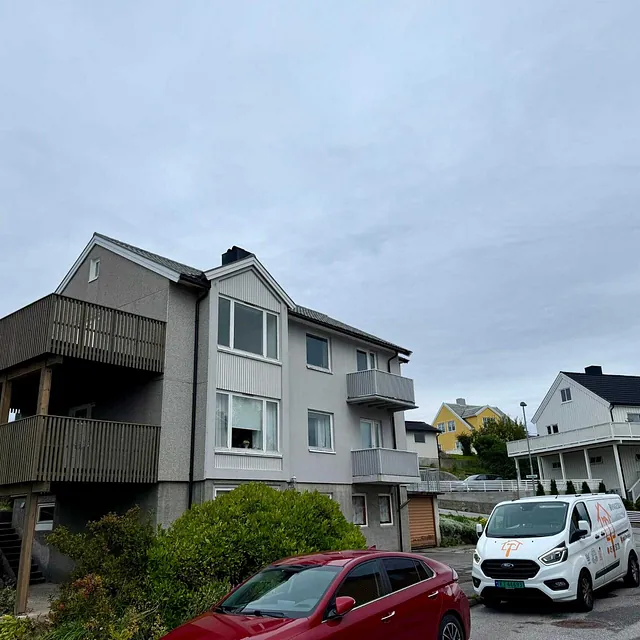 Eldre bolig med balkong og parkert bil i rolig nabolag under overskyet himmel.