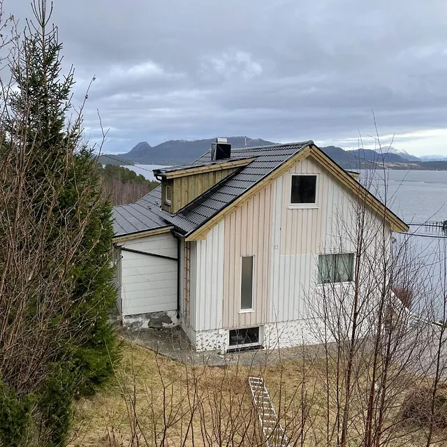 Hvit enebolig med svart tak, plassert ved sjøen og omgitt av natur.