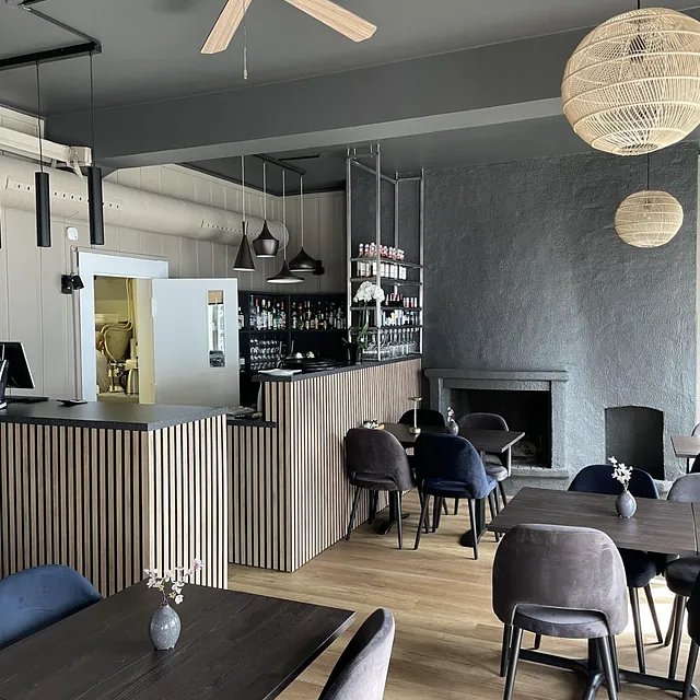 Bar og peis i restaurant med mørke bord, grå stoler og trelaminatgulv.