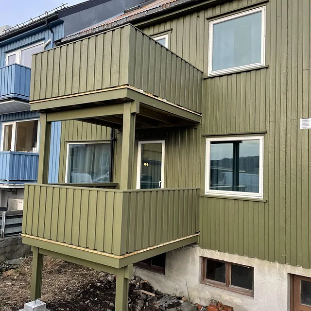 Grønnbemalt trehus med to balkonger og betongfundament.