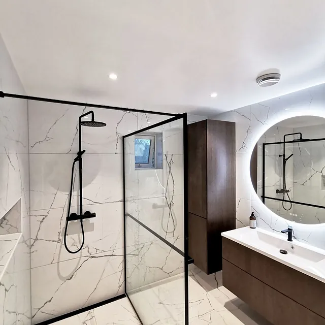 Bad med dusjhjørne i glass, mørke skap og rundt speil med LED-ramme.