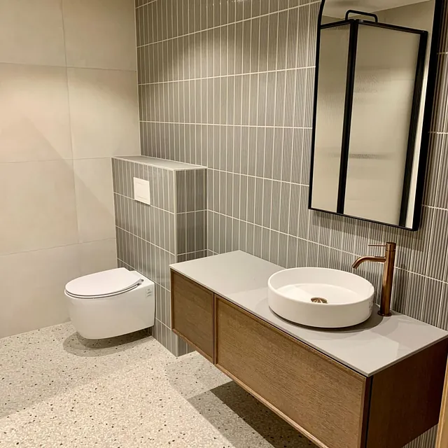 Bad med terrazzo-gulv, vegghengt toalett, servant med kobberkran og buet speil.