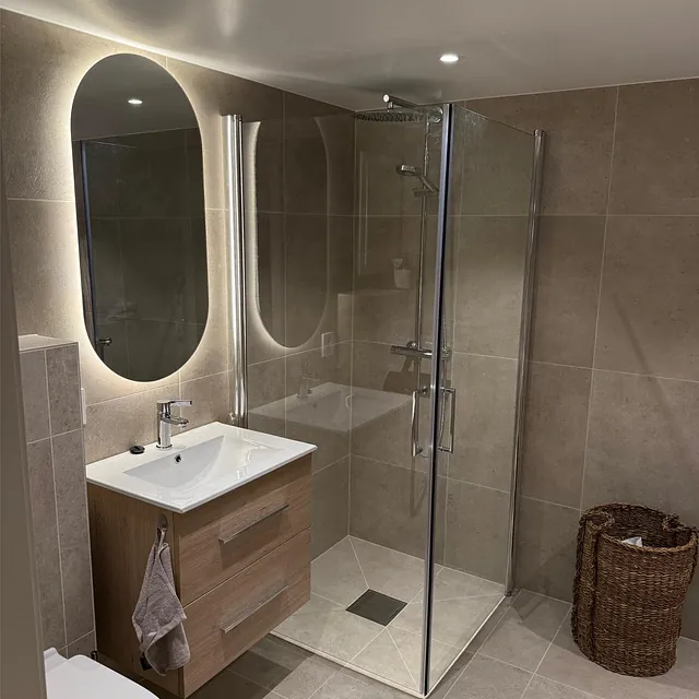 Stilrent bad med dusjhjørne, servant og speil med lys i varme nøytrale toner.